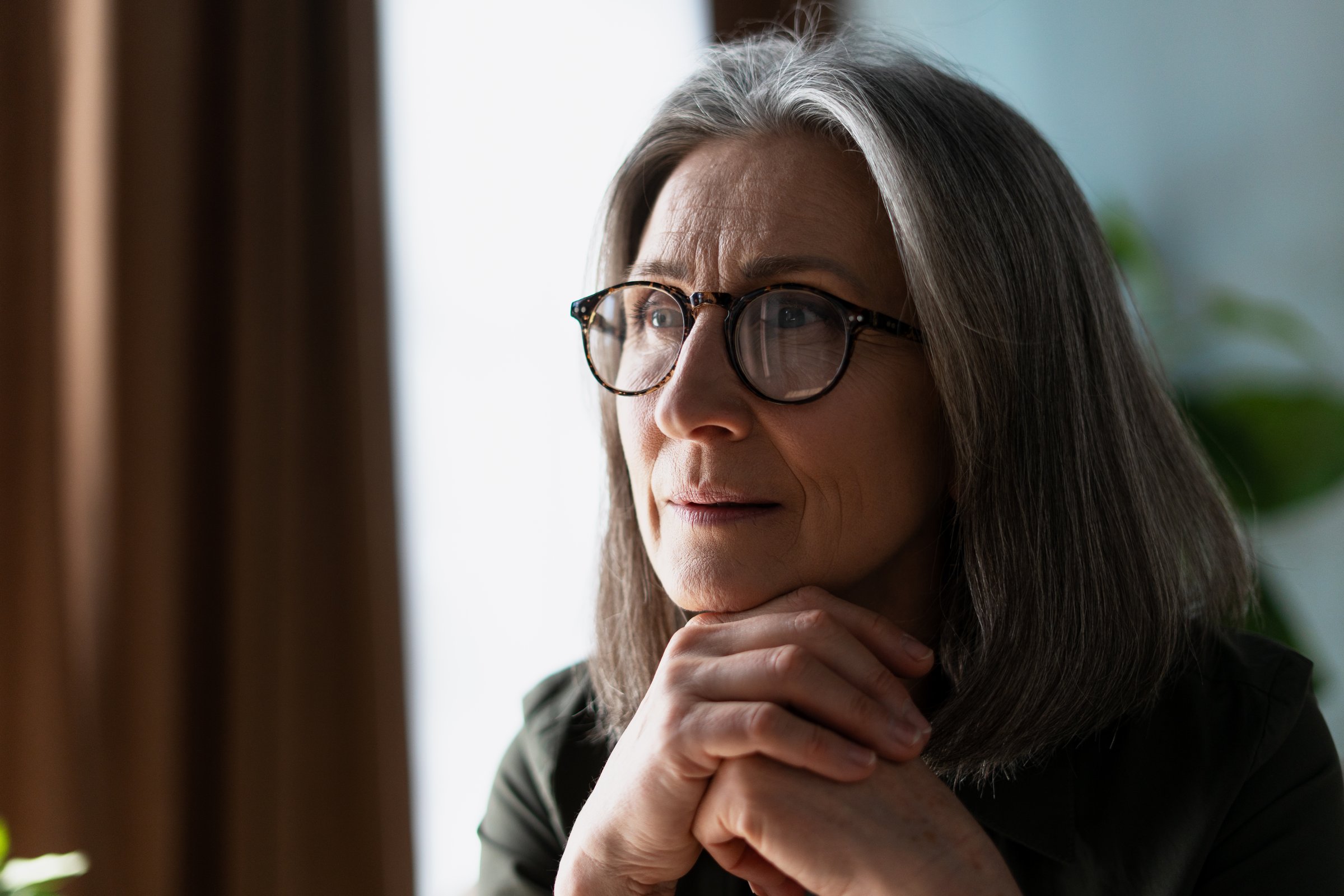 Thoughtful mature woman with glasses looking away, contemplating near the window at home or office