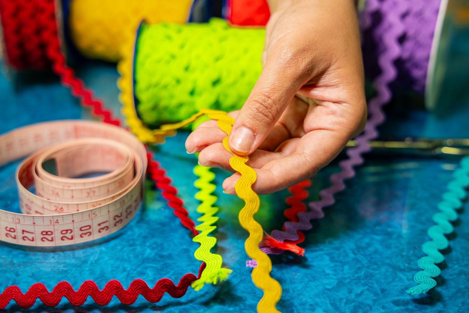 This close-up features a hand holding a textured yellow rickrack ribbon above a crafting surface filled with red, green, and purple trims. A measuring tape, scissors, and ribbon rolls create a vibrant DIY workspace scene focused on textile decoration and handmade projects