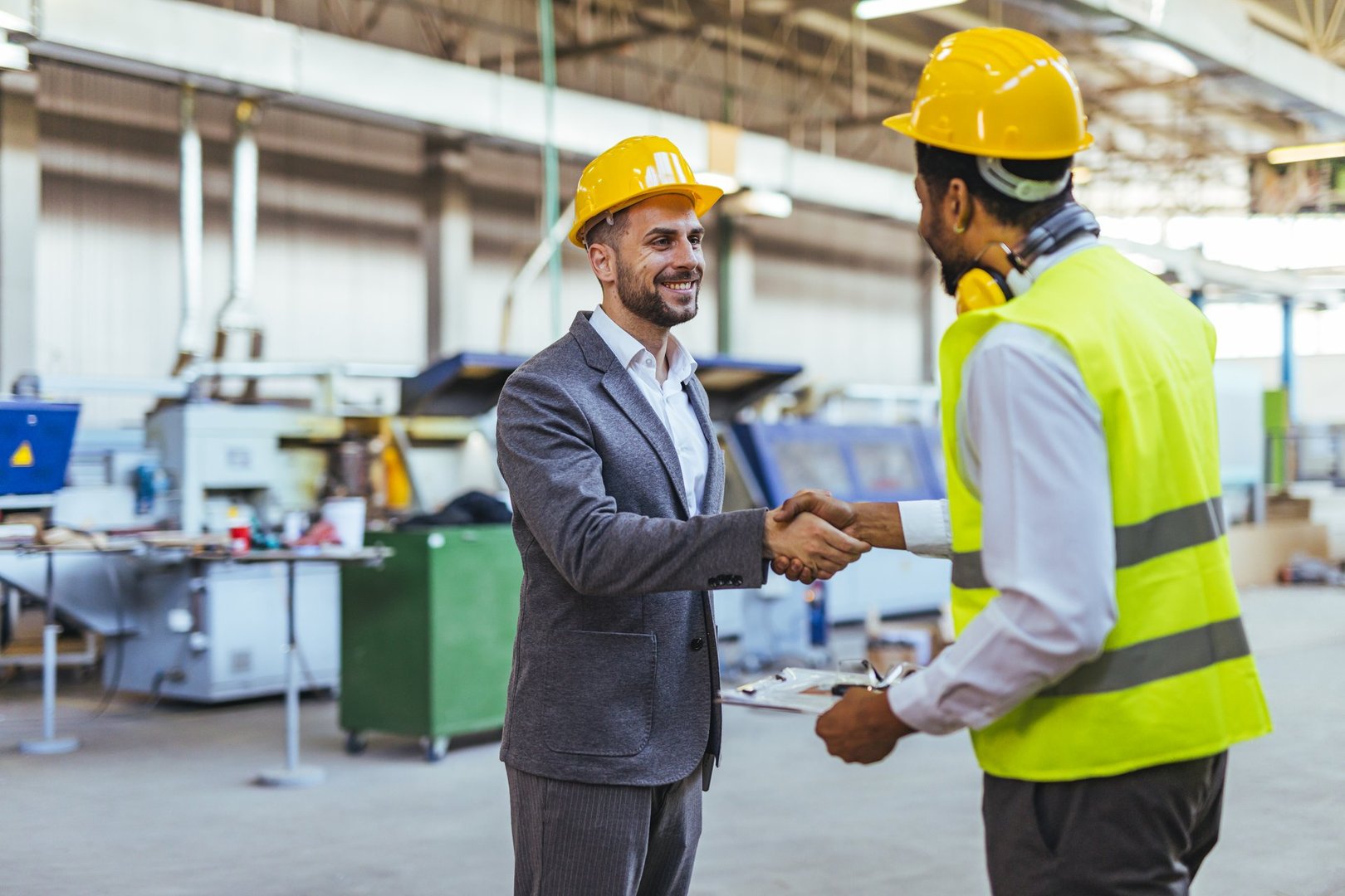 Two men wearing safety helmets and vests shake hands in a factory environment, signifying cooperation and teamwork. The machinery in the background highlights the industrial and production setting.