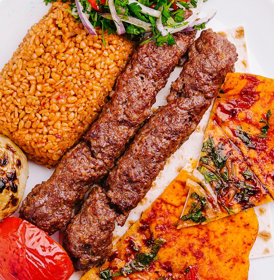A delicious plate showcases grilled kebabs accompanied by flavorful rice, fresh herbs, grilled vegetables, and a side of baked flatbread. This meal highlights rich textures and vibrant colors.