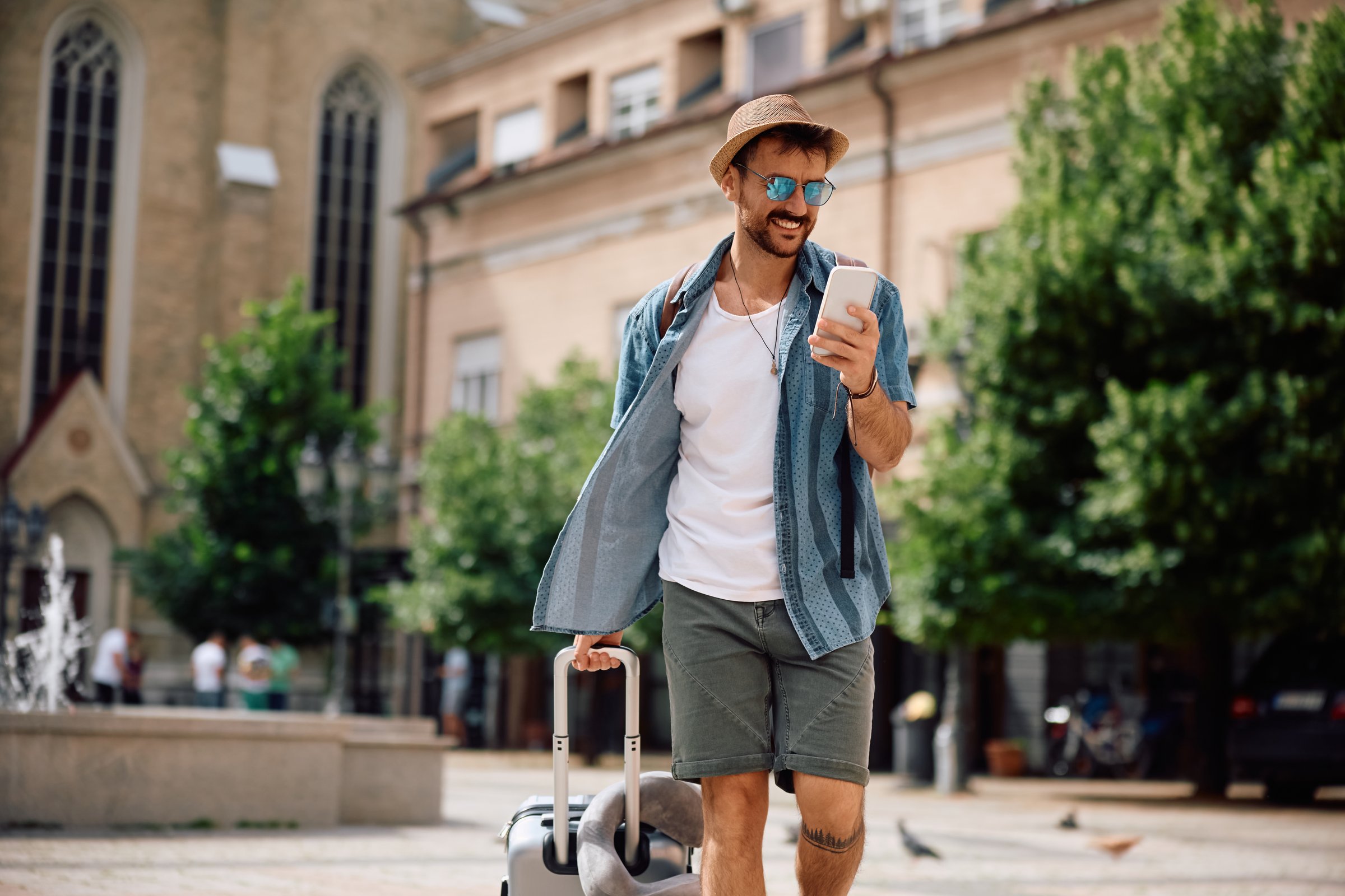 Happy traveler using mobile phone while walking with a suitcase on the street. Copy space.
