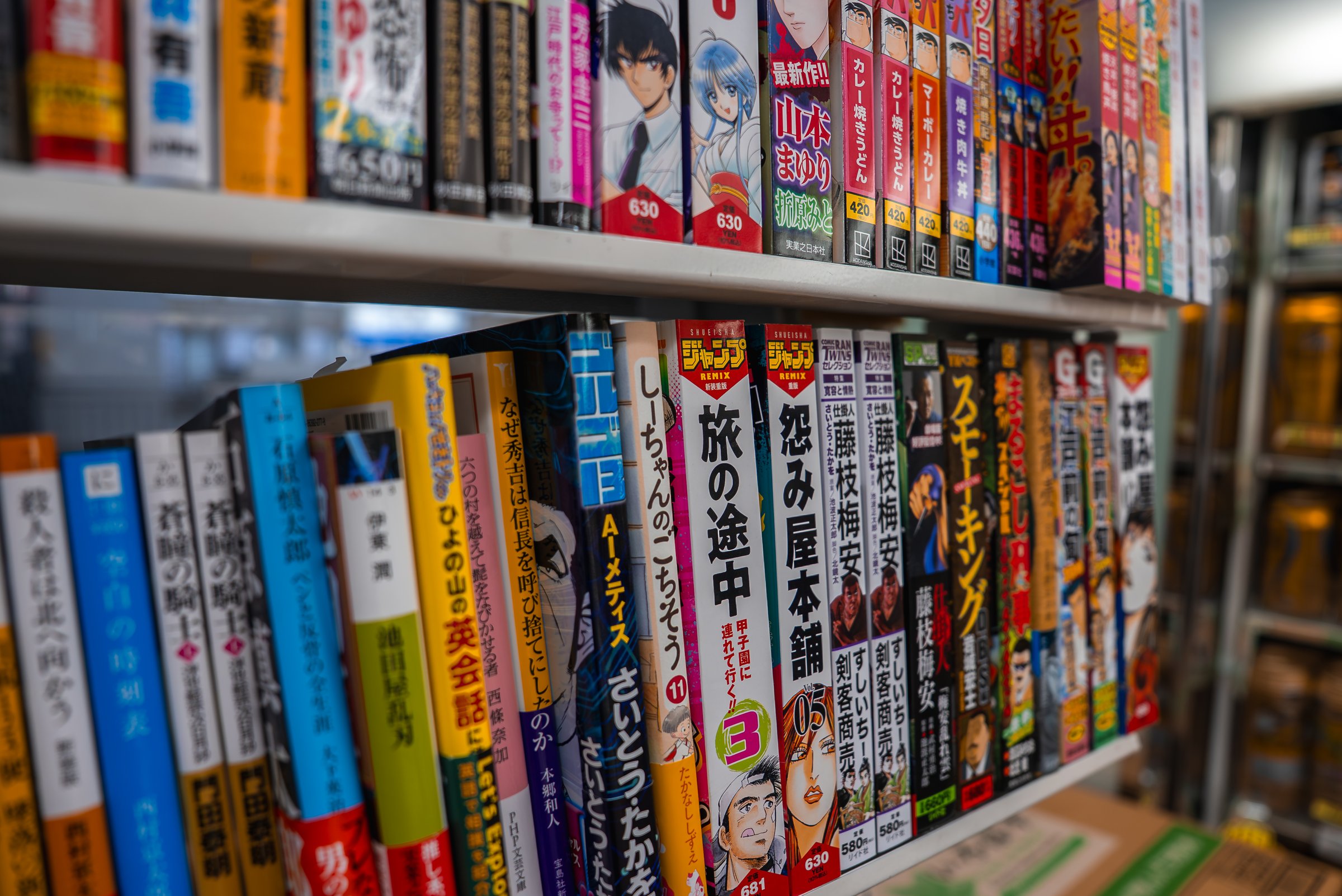 Rows of Japanese manga fill tidy shelves in an Osaka shop. Bold kanji titles and character art line colorful spines, eye level view, shallow depth, diagonal framing.
