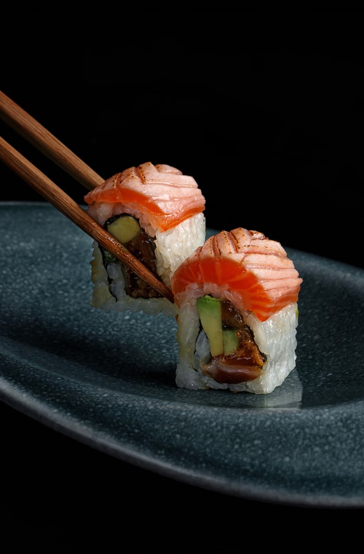Close-up of two sushi rolls filled with avocado, salmon, and other fresh ingredients, topped with microgreens, presented on a black background for a refined, minimalist look.