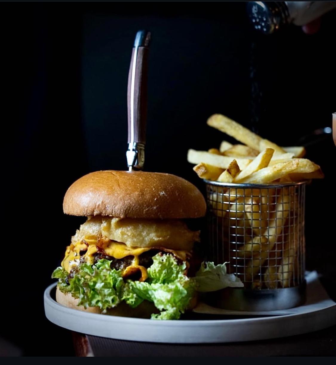 Cheeseburger with a knife on top, side of fries in a metal basket, dimly lit background.