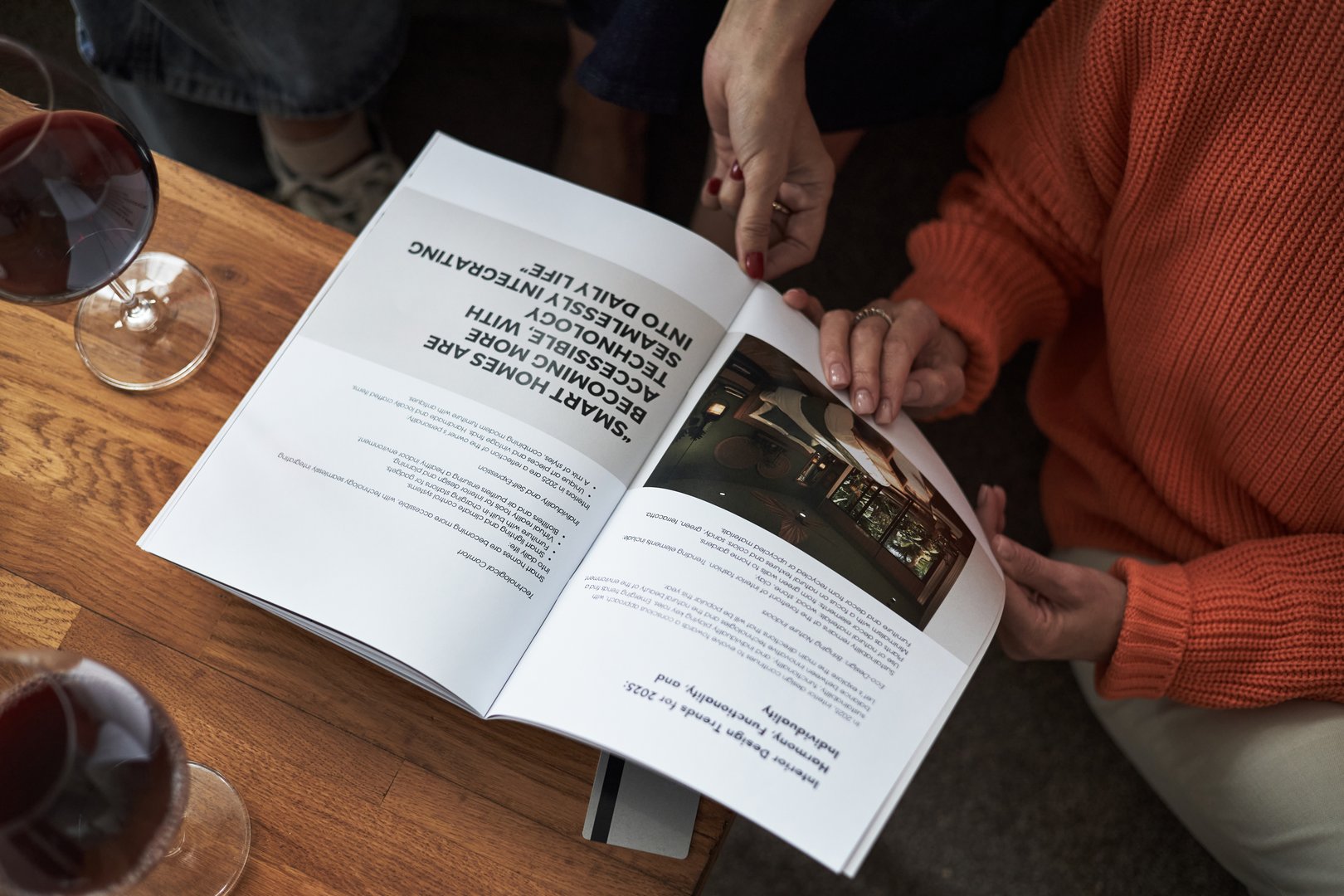 Two young adult Caucasian women sitting together reading open magazine on wooden table, hands visible holding pages, glasses of red wine nearby