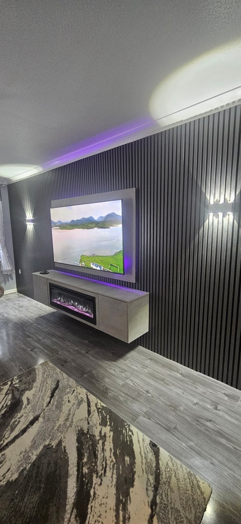 Modern living room with a wall-mounted flat-screen TV, black slatted wall, electric fireplace, and ambient lighting.