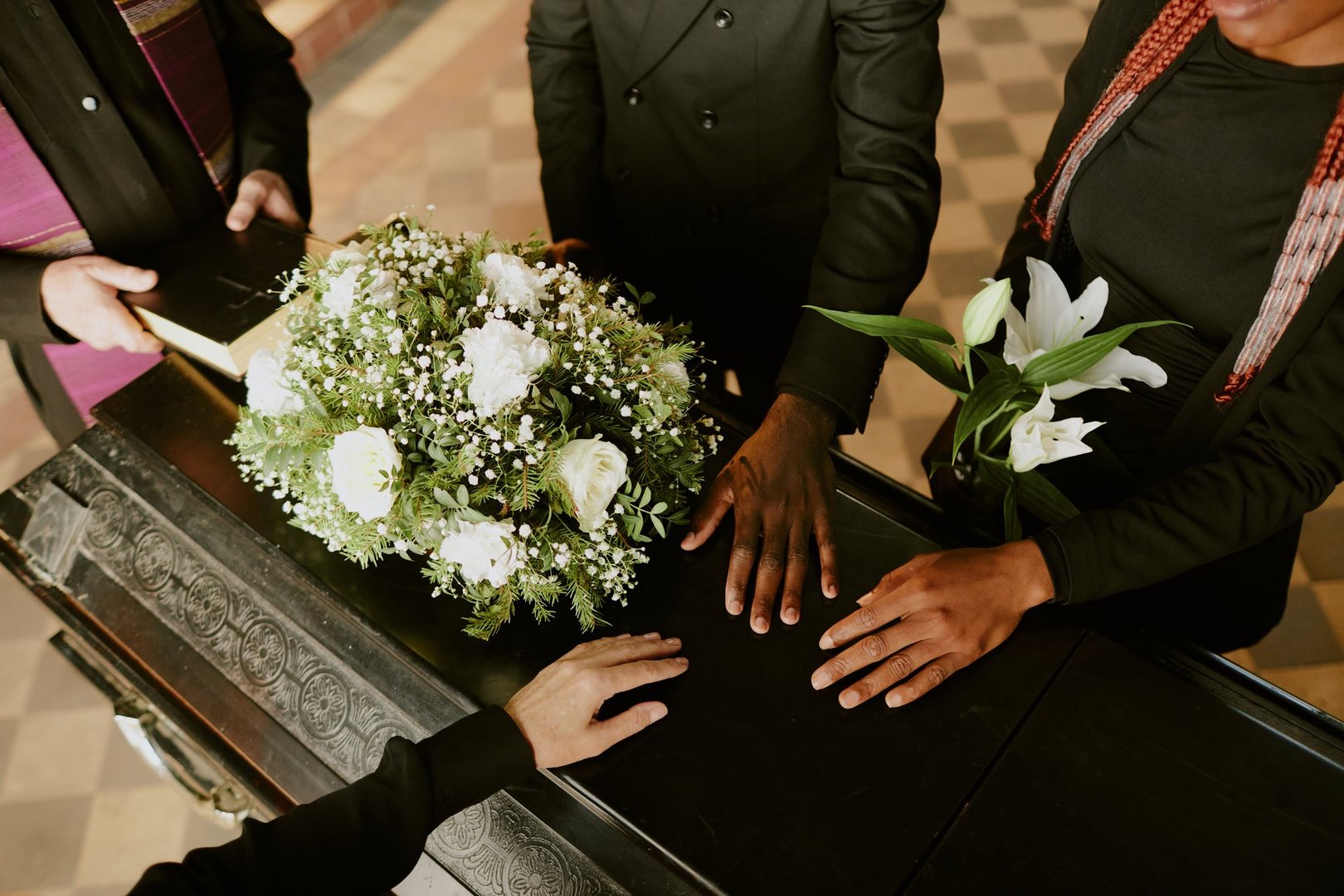 High angle shot of unrecognizable hands on black wooden coffin with white roses on top, pastor holding holy book