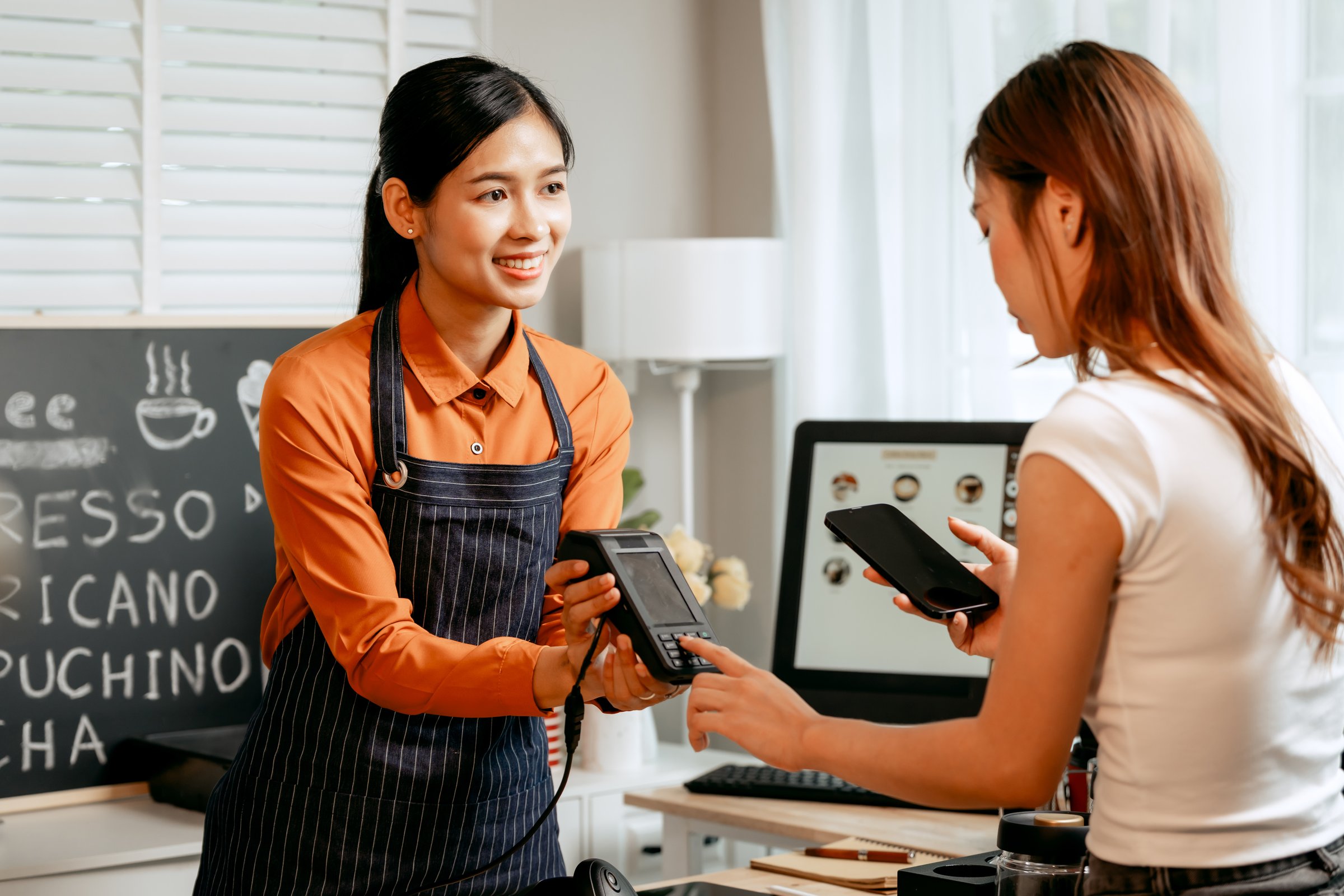 A young Asian female barista in modern cafe assists woman customer ordering latte, fresh coffee beans, dark roast, croissant, completing the purchase with mobile payment at the POS using QR code.