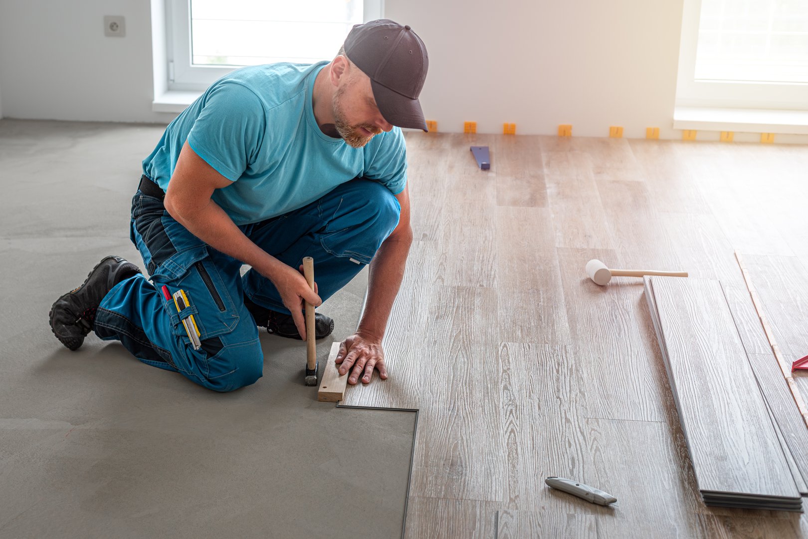 Floating floor work. The worker taps the board of vinyl plank to lock the click system. He is using hammer and wooden plank.