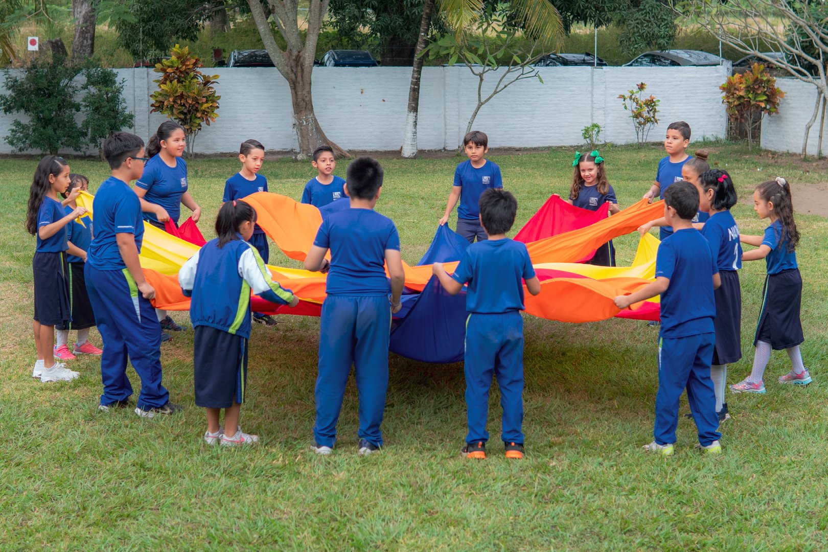 Children in blue uniforms playing with a colorful parachute outdoors on grass, forming a circle.