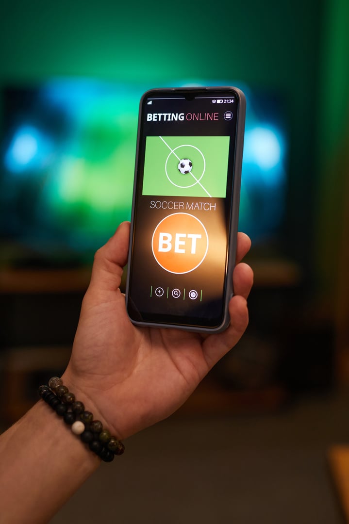 Hand holding smartphone showing a soccer betting app interface with live match. Device illuminates with screen's bright display