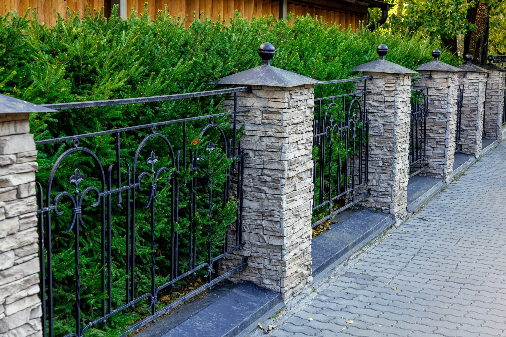 A stone and metal fence enhances the beauty of greenery along a brick pathway in a suburban neighborhood