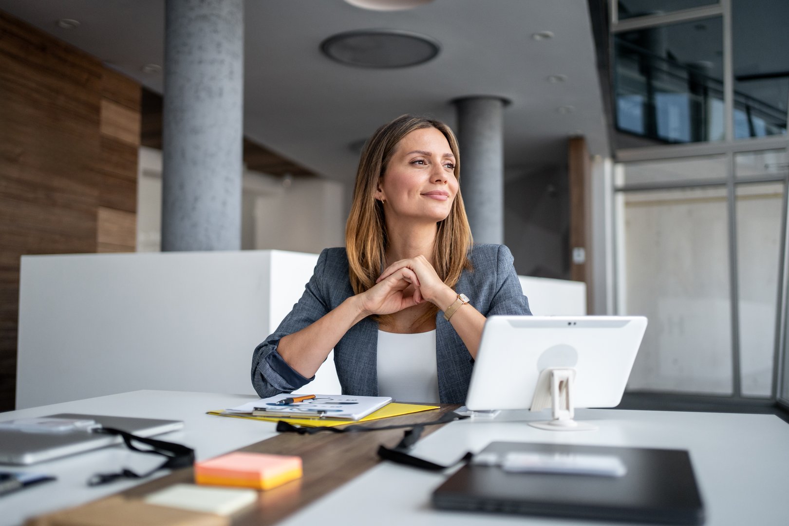 Businesswoman sitting at a modern office desk, smiling and gazing thoughtfully into the distance while contemplating future business plans and aspirations for success