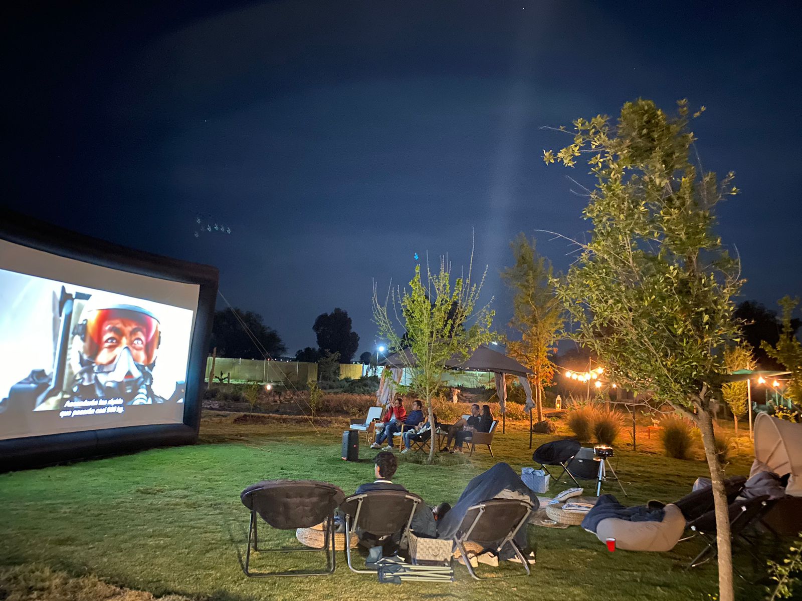 People watching a movie outdoors at night on an inflatable screen, surrounded by trees and seating on a grassy area.