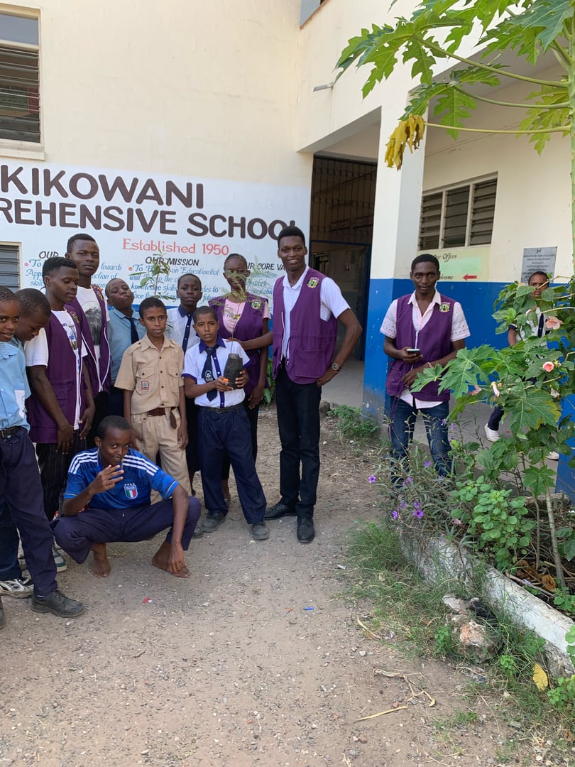Group of students and staff outside Kikowani Comprehensive School building, two wearing vests, with one seated on the ground.