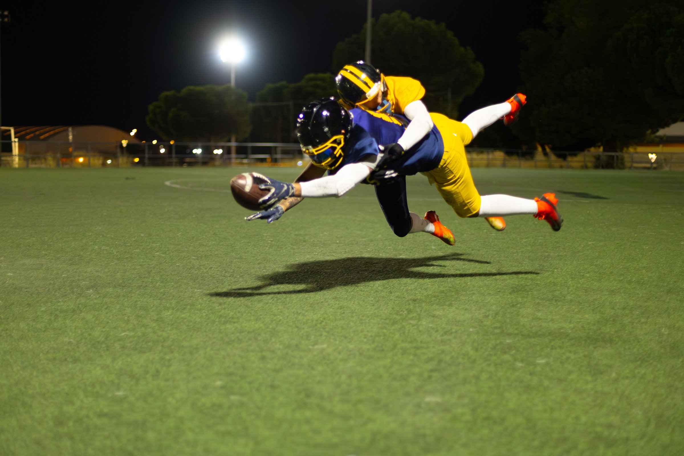 American football players in helmets and uniforms diving for the ball in action on the field during a competitive night game
