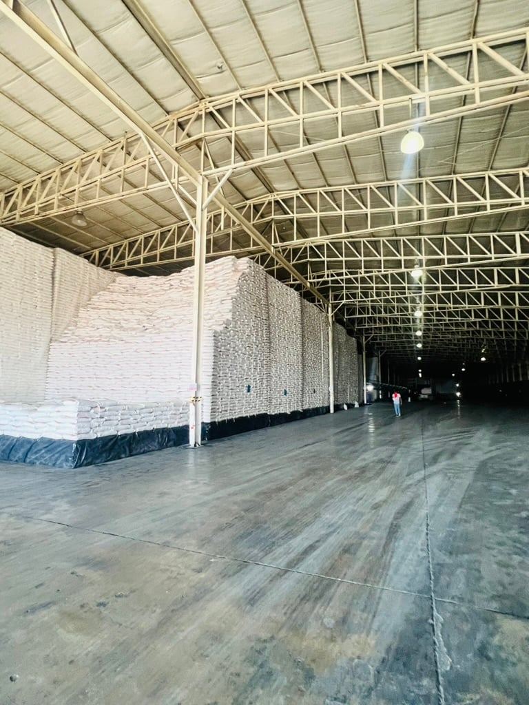 Large warehouse interior with stacks of white bags piled against the high walls under a metal roof.