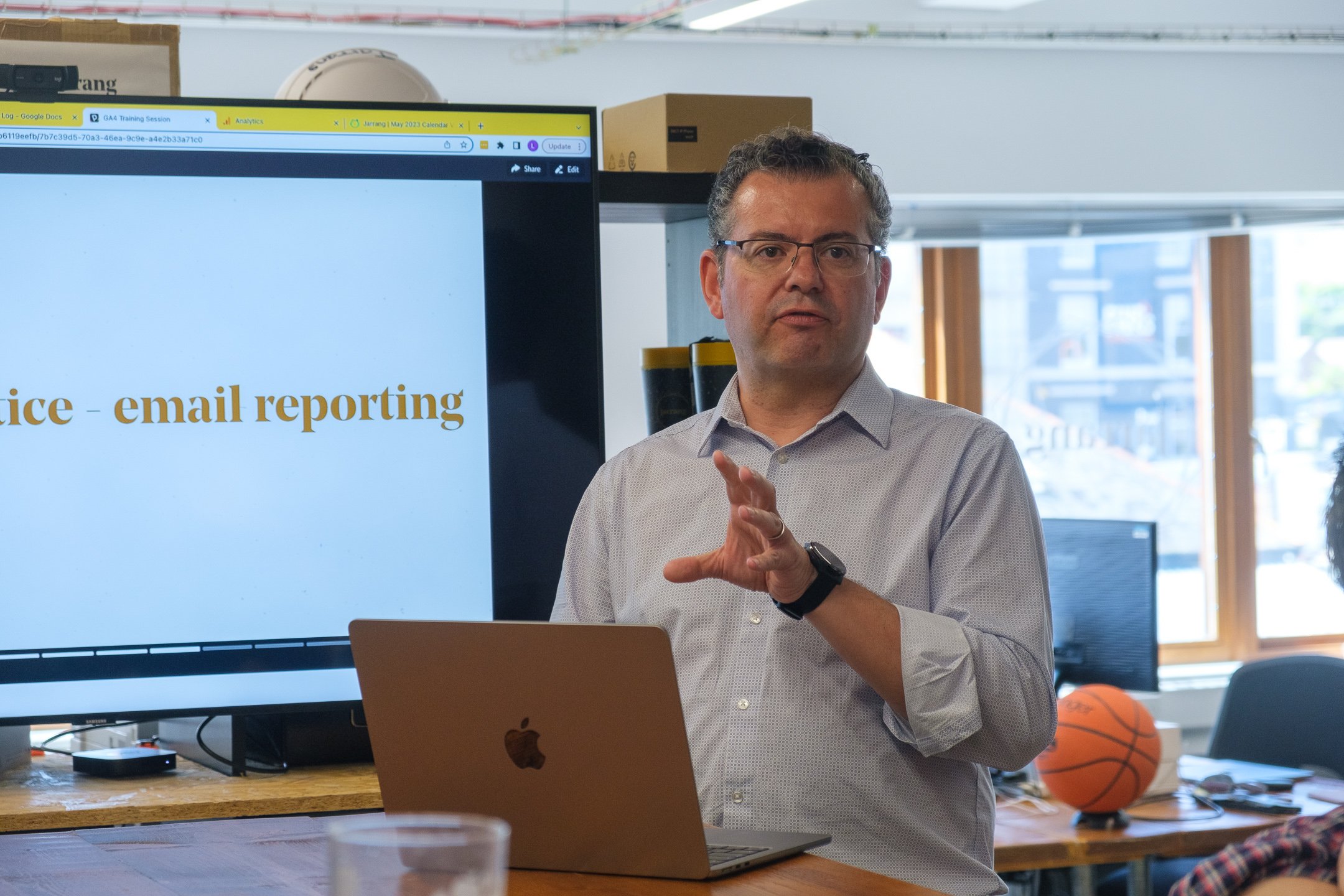 A man giving a presentation in an office with a laptop and screen displaying office - email reporting.