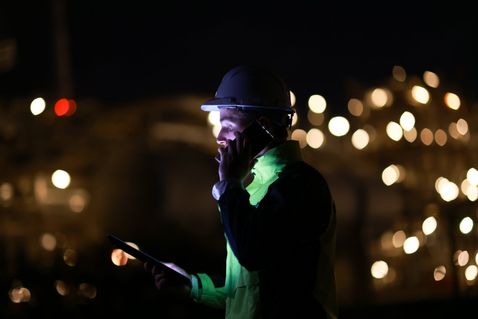 Smart  engineer with green safety jacket works on a digital tablet and using a mobile phone at the refinery plant during night shift