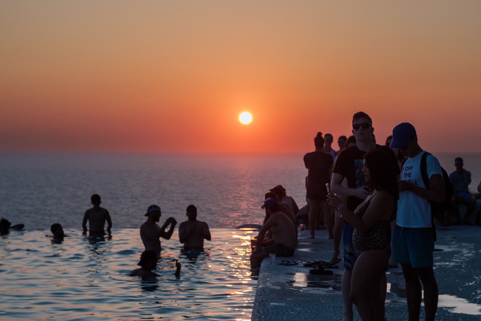 Ios, Greece - July 31, 2019 : Young people gather around a swimming pool, overlooking the Aegean sea, to watch the sunset in Ios Greece