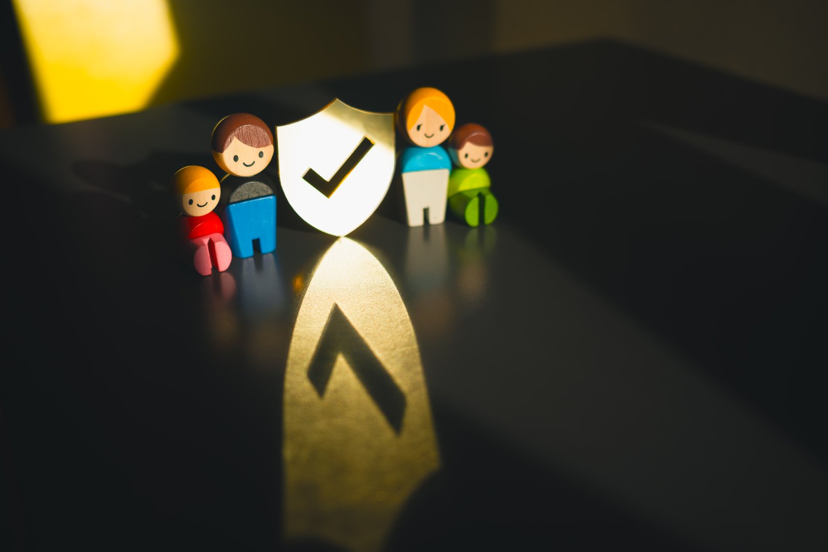 Wooden family figures with shield and checkmark symbolizing family protection, home security, life insurance, safety assurance, and caring support for parents and children in a trusted environment.