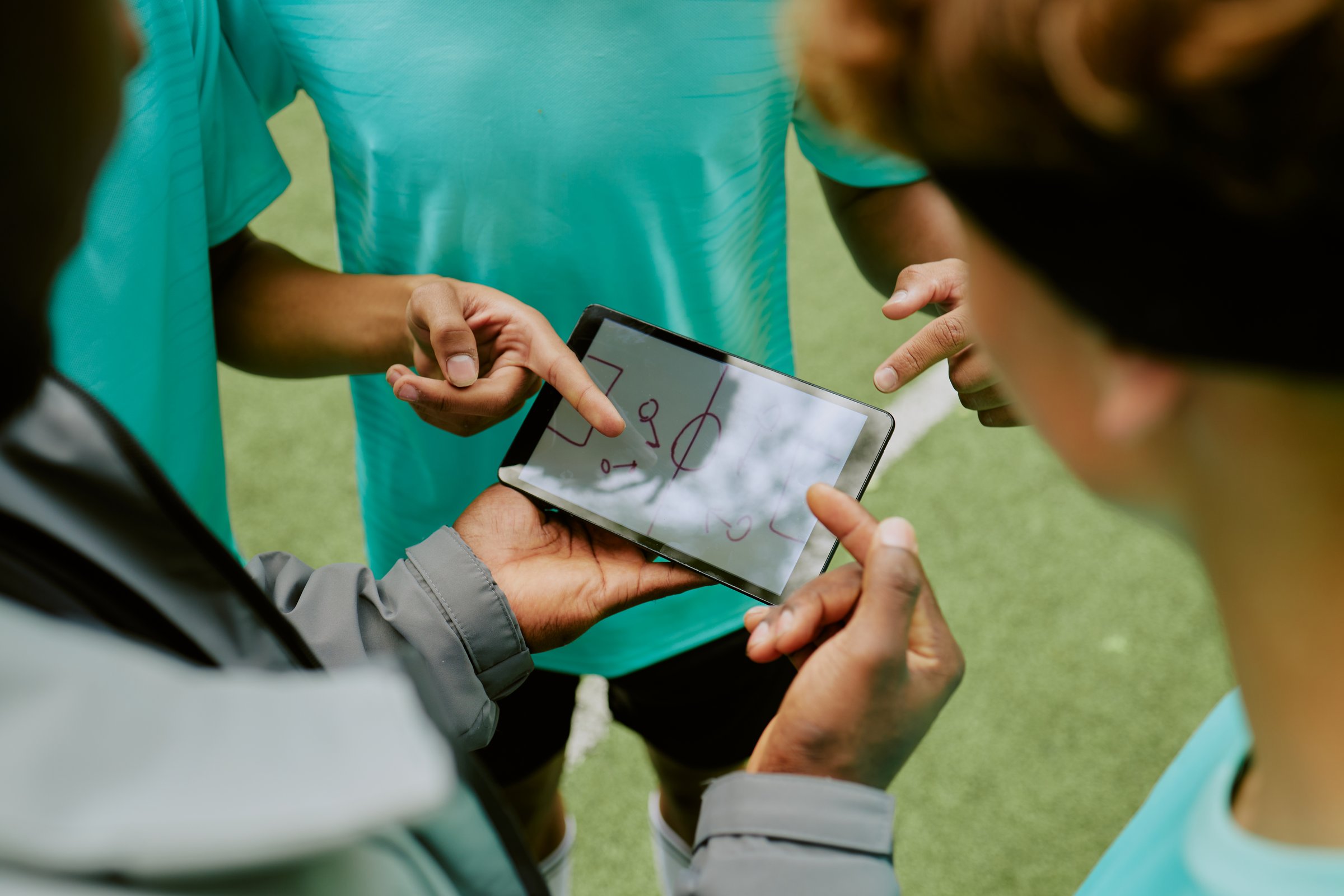 Group of teenage boys discussing sports strategy while pointing at digital tablet displaying play diagram outdoors, focusing on teamwork and planning during practice session