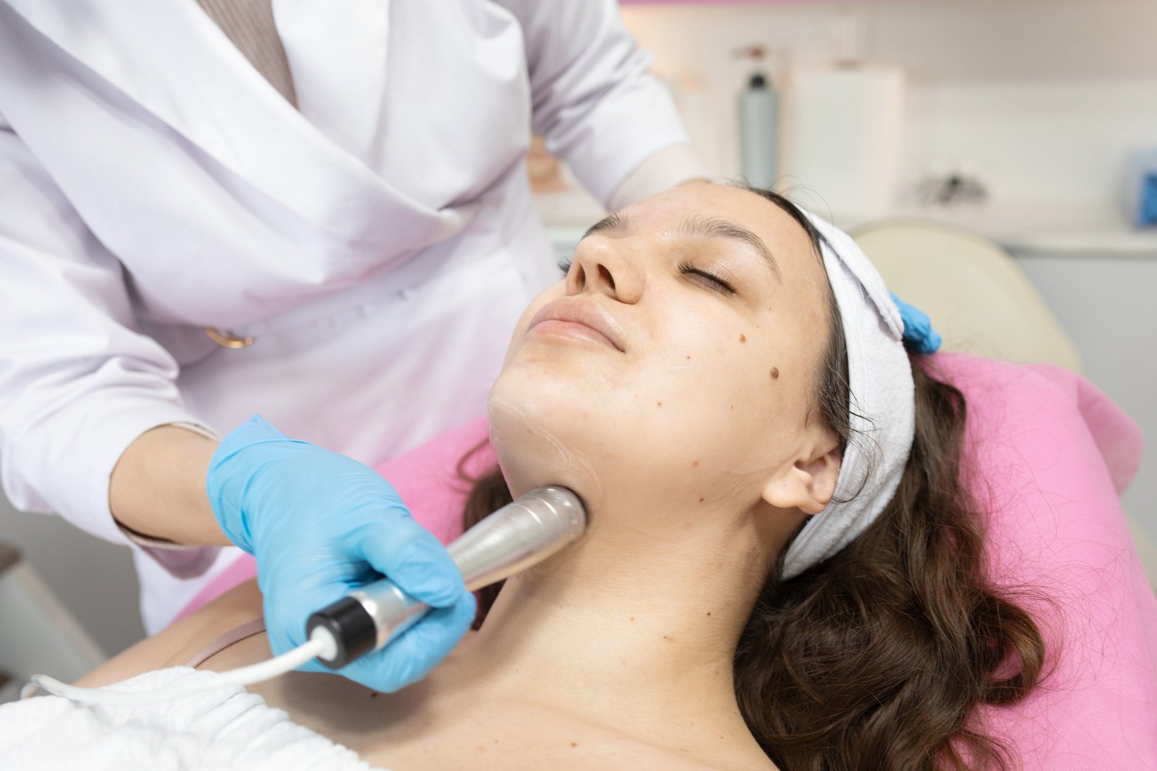 Woman is getting a facial treatment with a rf lifting device on her face. The woman is wearing a white towel and a blue glove