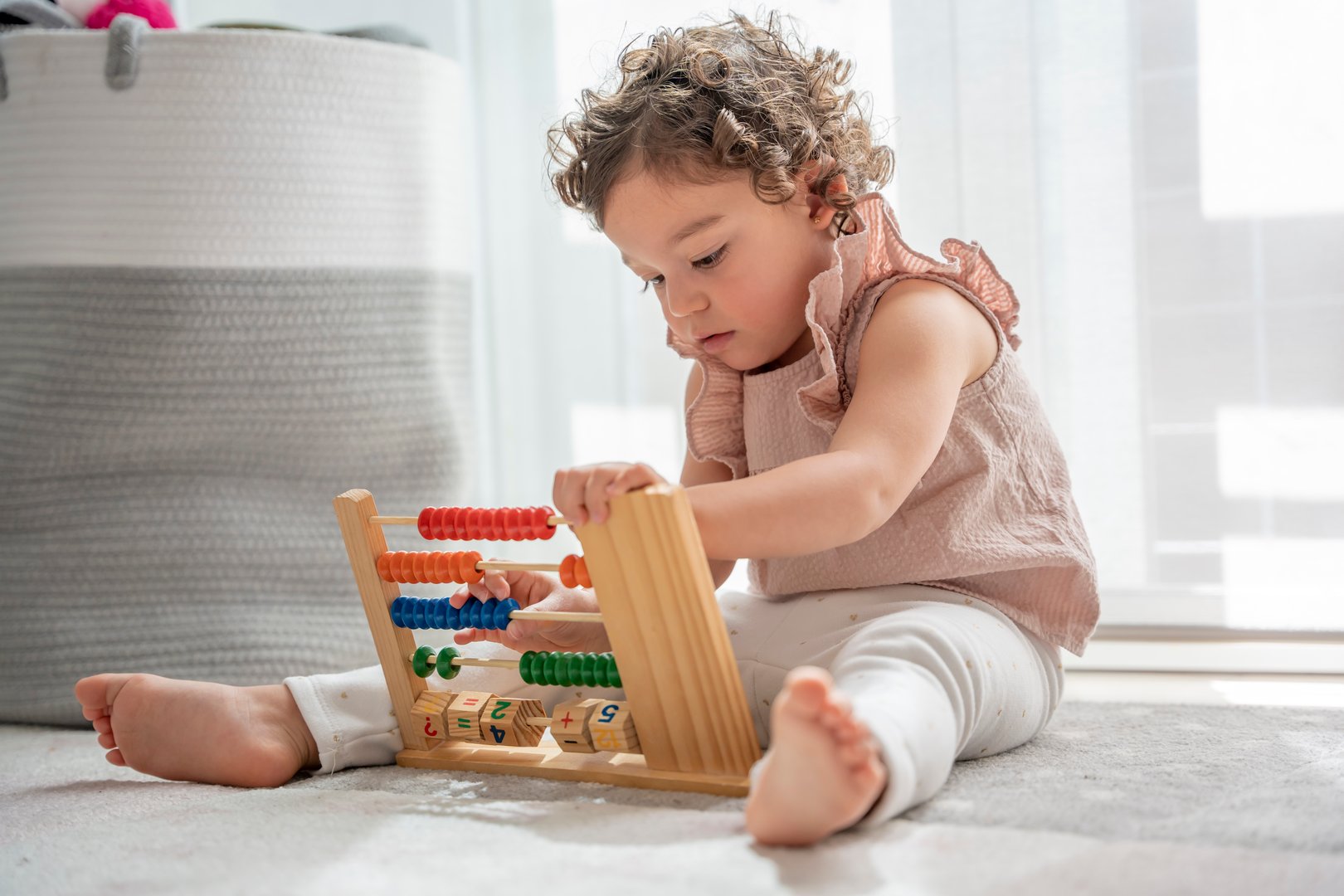 little girl with curly hair playing sitting on the carpet in her room with a wooden toy made of beads, numbers and colors. montessori material concept development and learning in the childhood stage.