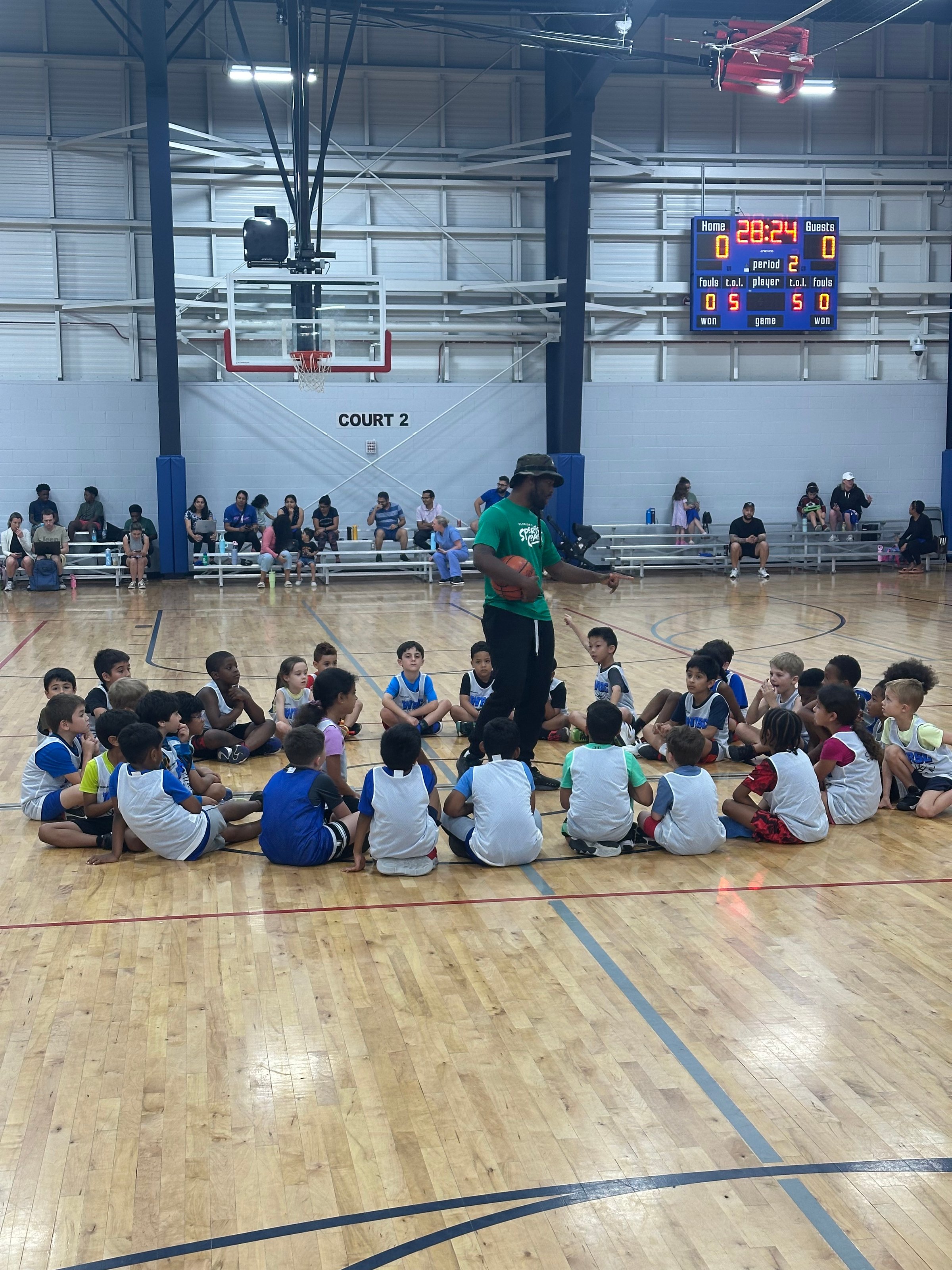 Coach instructing young basketball players sitting on a gym floor, with parents watching from the sidelines.