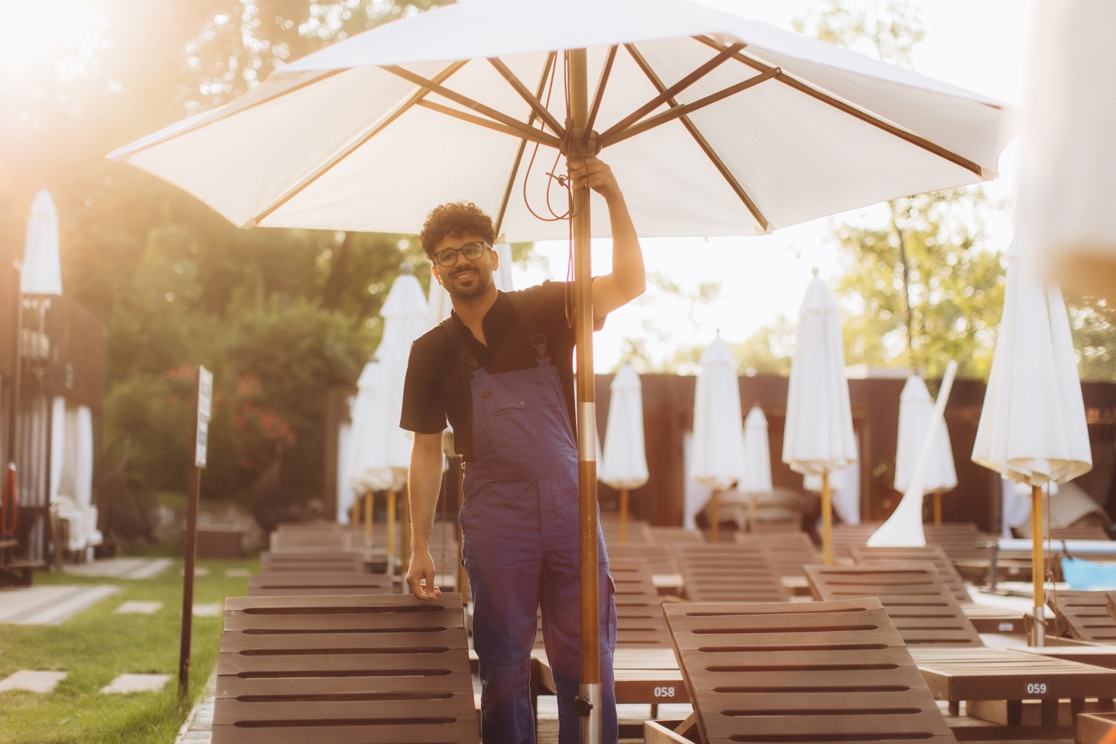Maintenance worker setting up white beach umbrellas and wooden chaise lounges at a poolside resort, preparing for the summer season