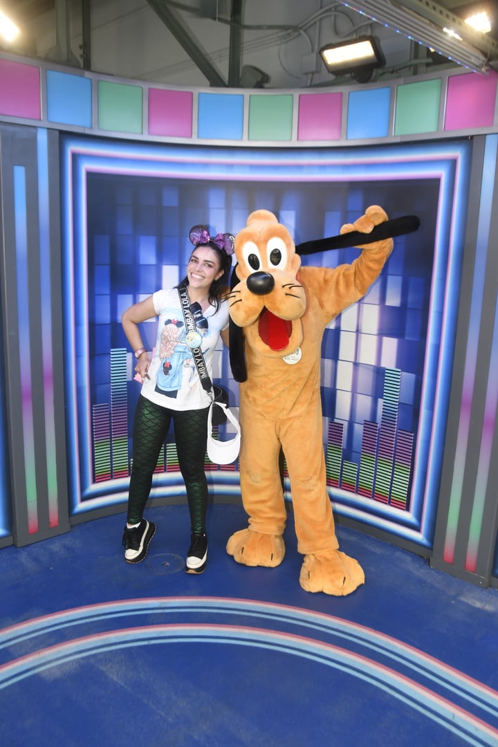 Person wearing Mickey ears posing with Pluto mascot in a colorful backdrop with soundwave graphics.