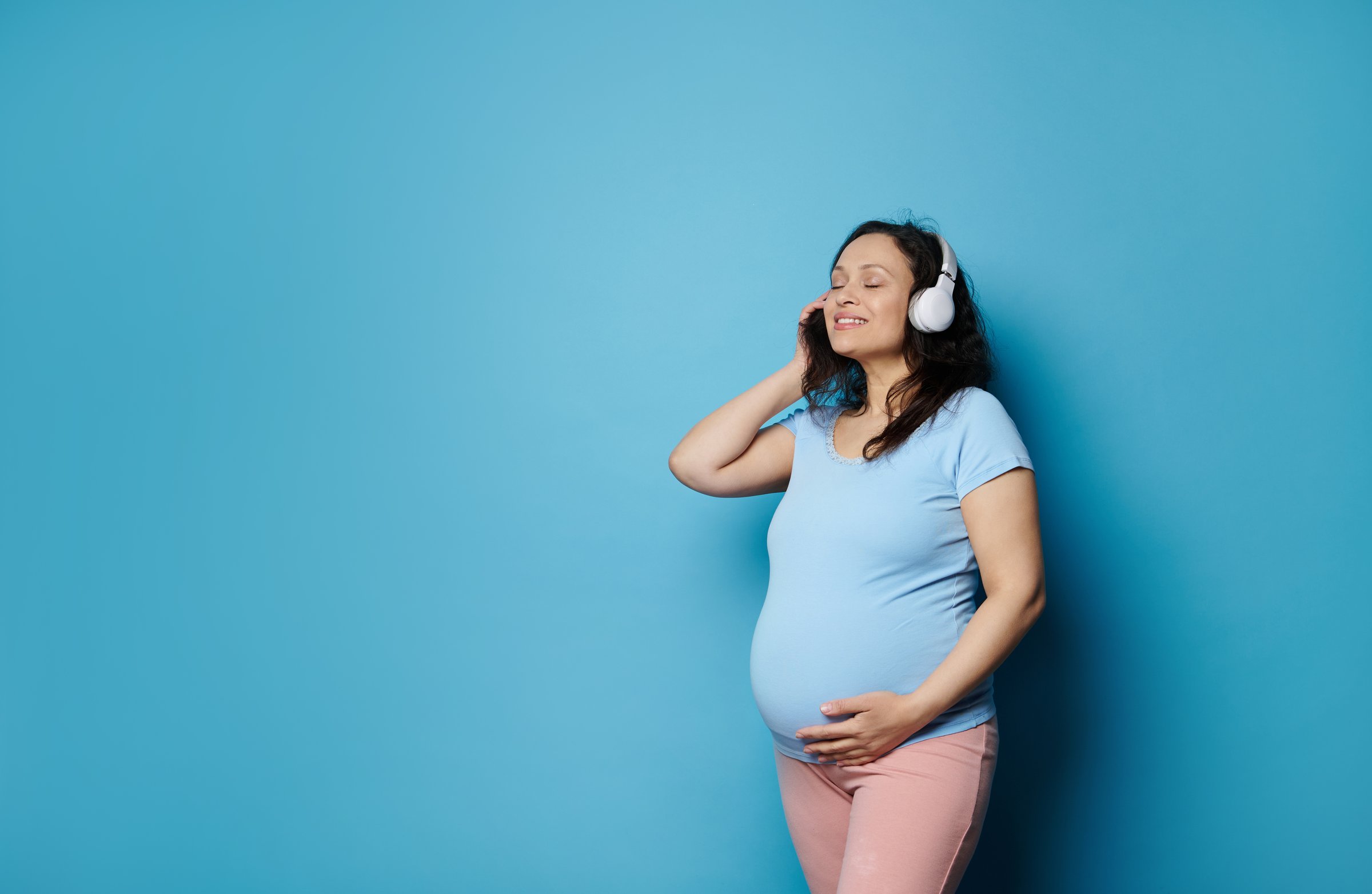 Happy smiling adult pregnant woman, expectant mother enjoying soundtrack on white headphones, stroking her belly, expressing positive emotions from pregnancy and maternity, isolated on blue background