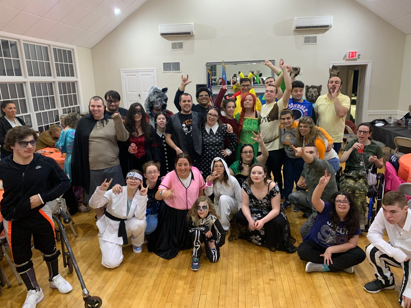 Group of people in Halloween costumes posing in a brightly lit room, some wearing face makeup and masks.