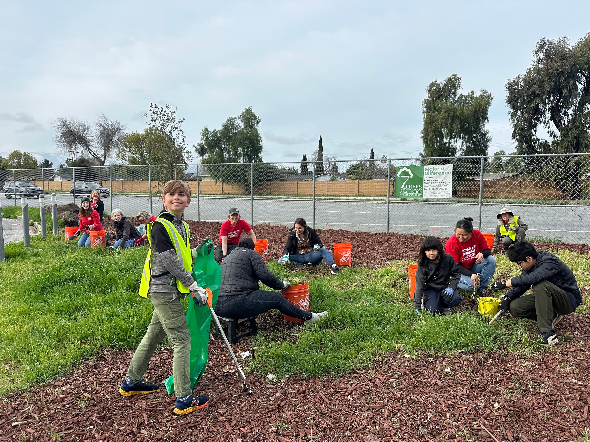 Volunteers in safety vests and gloves planting and cleaning a garden area beside a road, using buckets and tools.