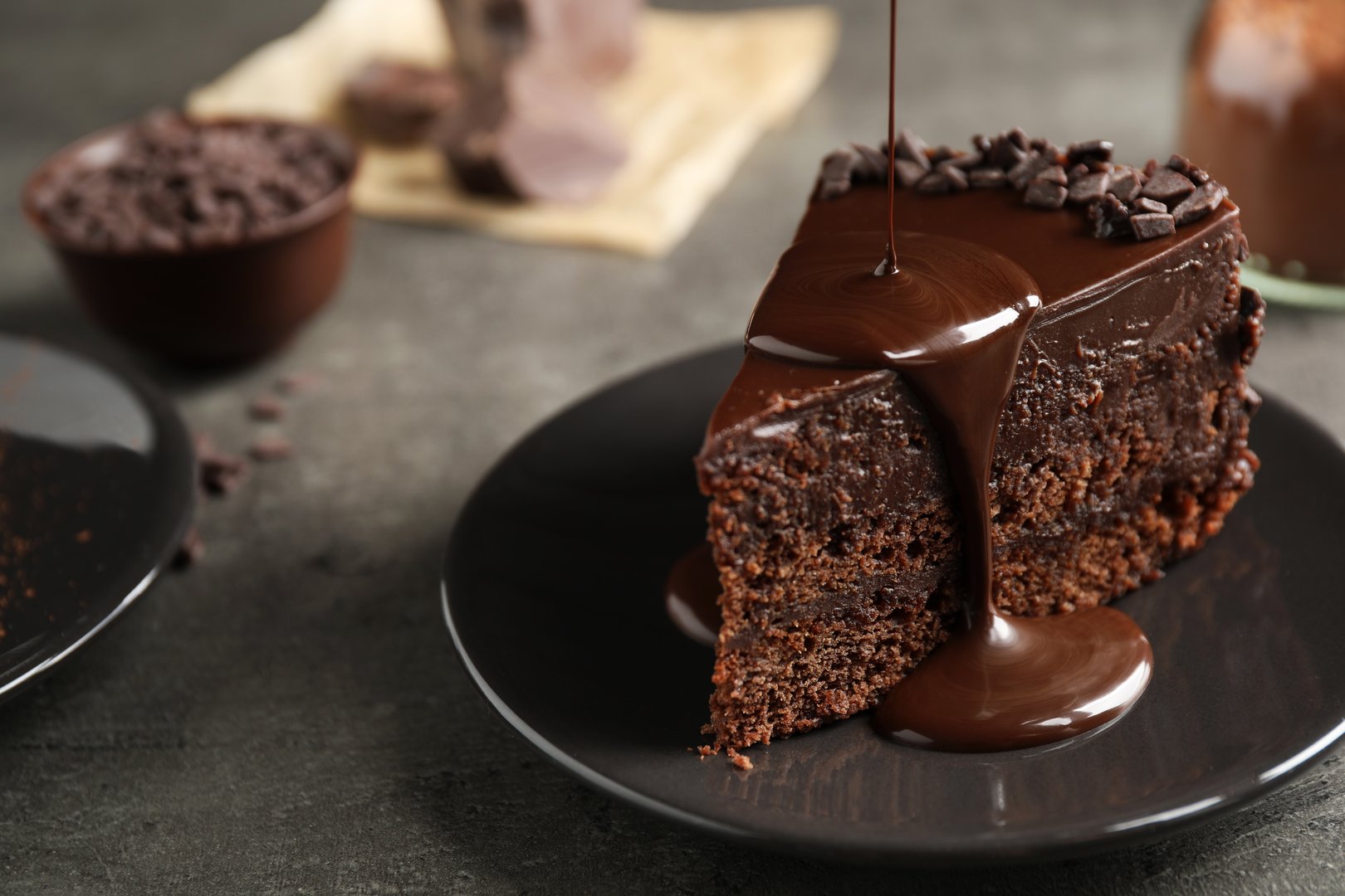 Close-up photograph of a moist, layered chocolate cake slice resting on a dark ceramic plate, featuring a thick, smooth stream of warm chocolate sauce pouring over the top. This captures indulgence, dessert luxury, and sweet confectionery, suitable for bakery menus and food advertisements.
