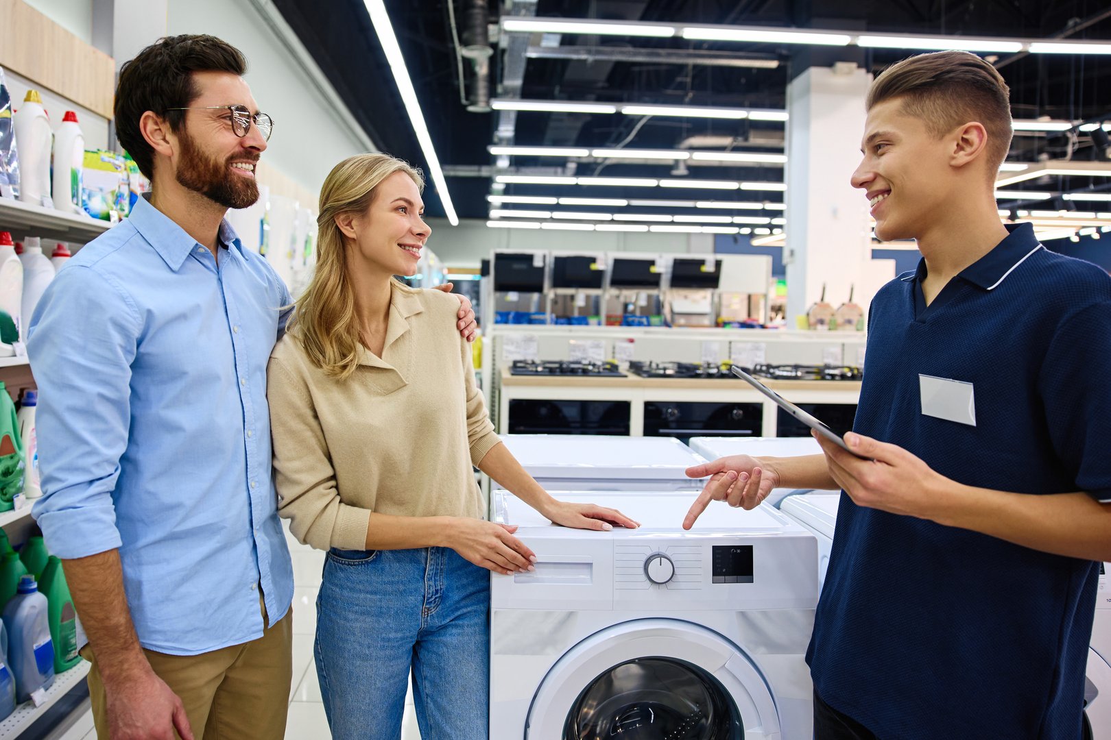 A couple engages in conversation and interaction with a knowledgeable salesperson while actively exploring and examining a variety of washing machines at a contemporary electronics retail store