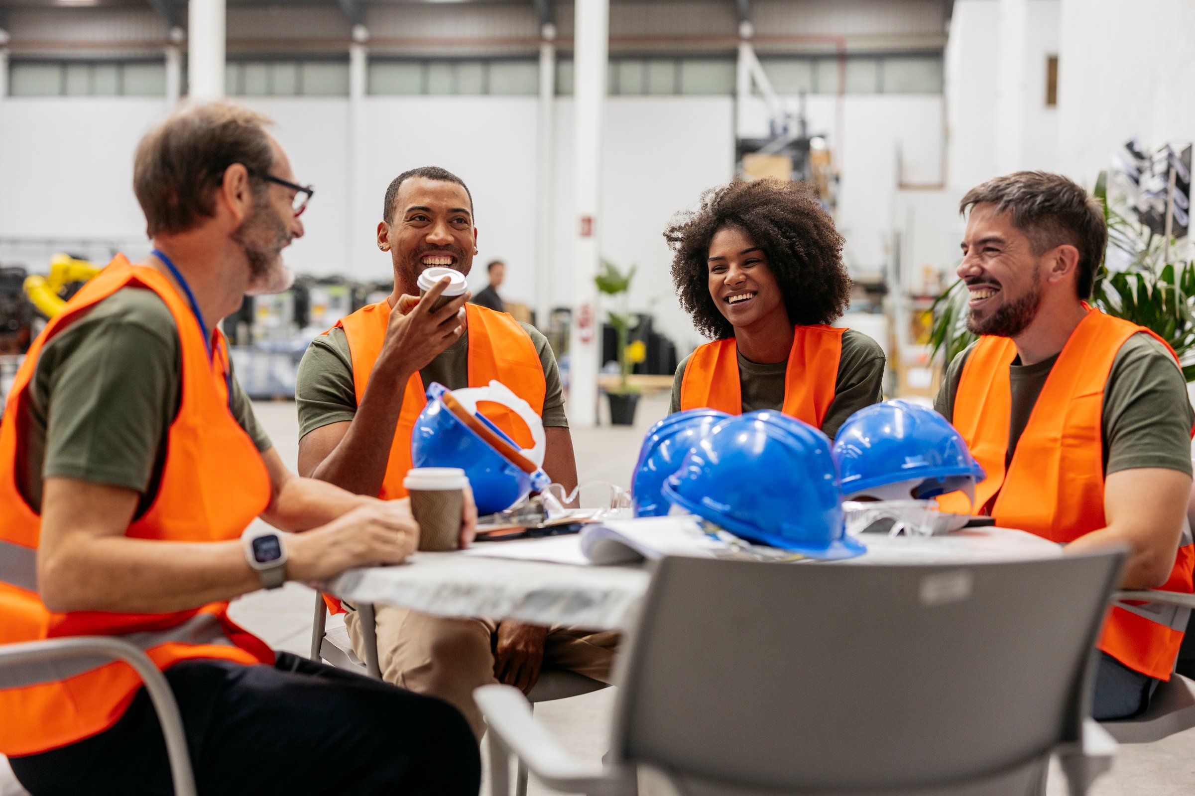 Four smiling multiracial engineers wearing orange safety vests are enjoying a coffee break at a round table in a factory, with blue safety helmets placed on the table