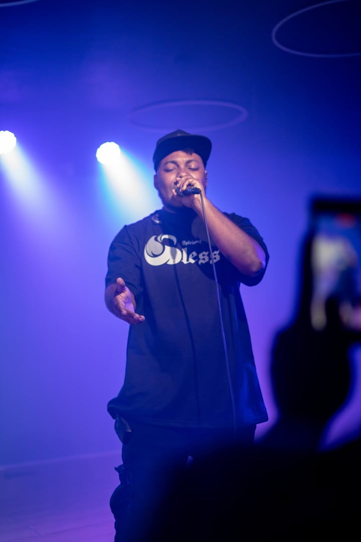 A man performs on stage with a microphone, wearing a black cap and t-shirt, against a blue-lit background.