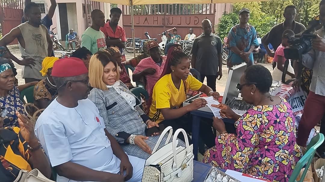 People seated outdoors participating in a community health registration event. A woman in yellow assists.