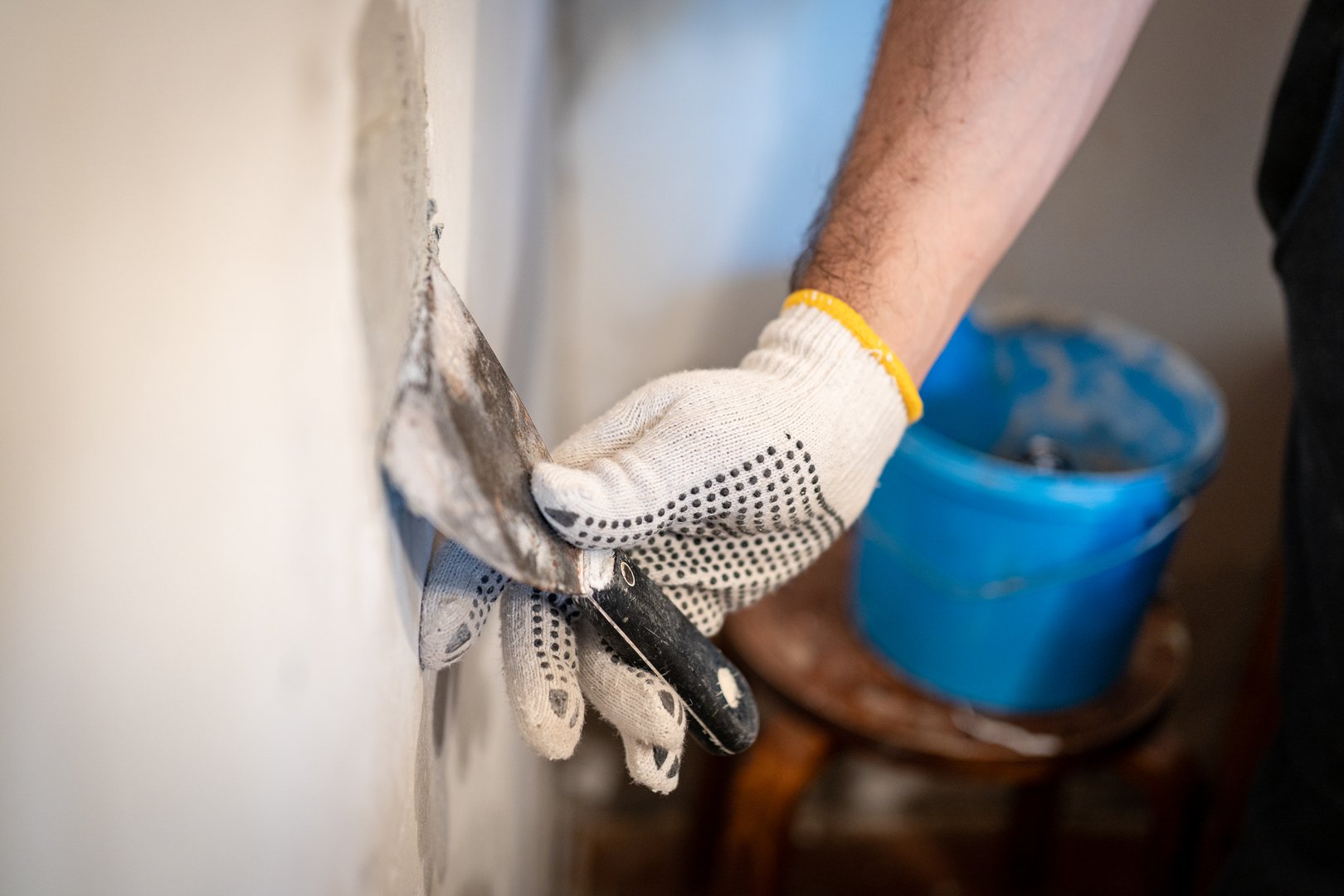Close-up of a worker's gloved hand using a small putty knife to press thick patching compound firmly into a hole in the interior wall.