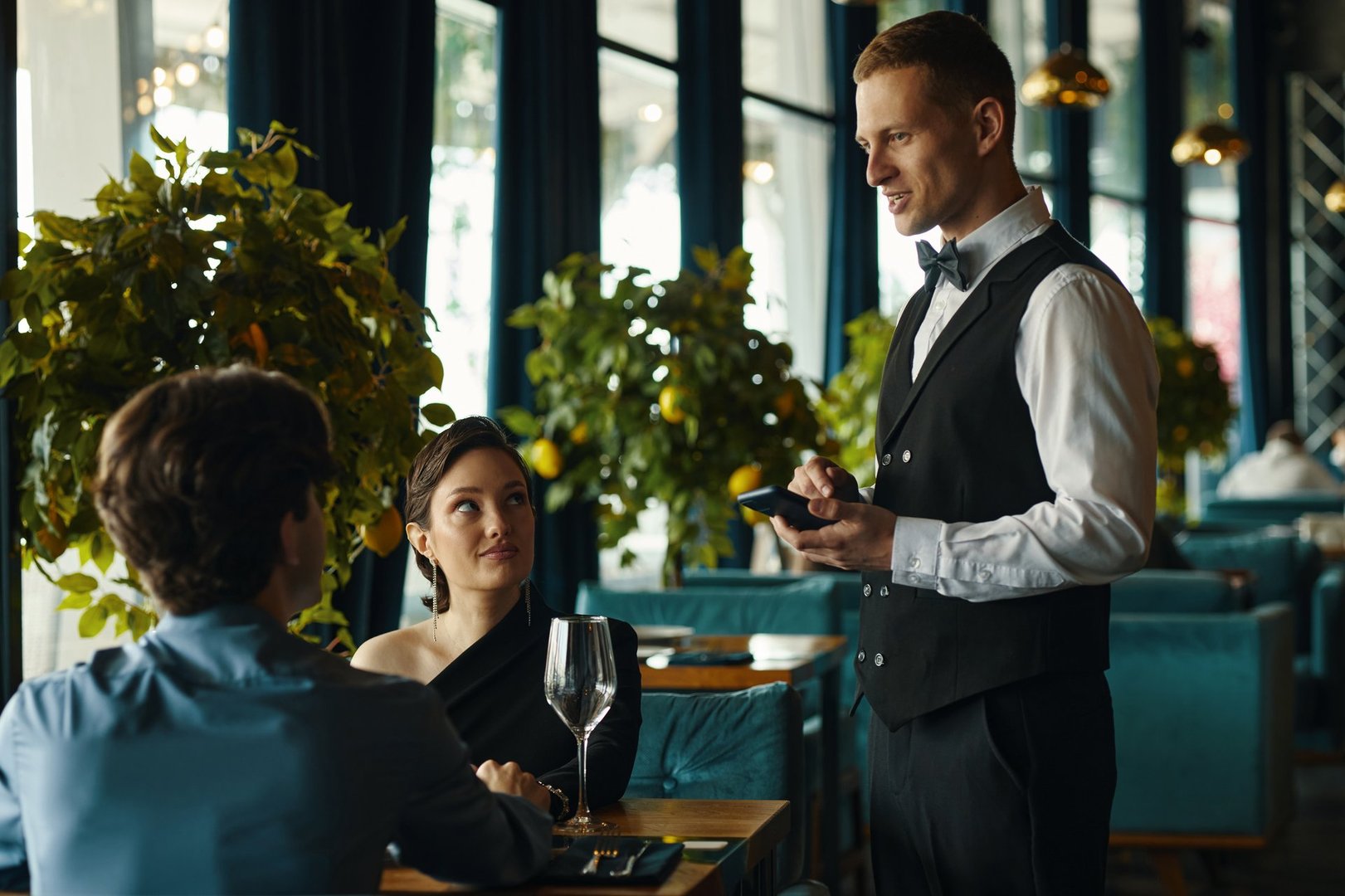 Caucasian young adult male waiter taking order from young adult Caucasian man and young adult Asian woman sitting at restaurant table, woman looking at waiter, man facing away