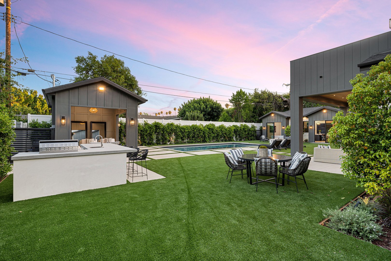 A modern backyard with a swimming pool, outdoor seating, and a small building under a pink and blue evening sky.