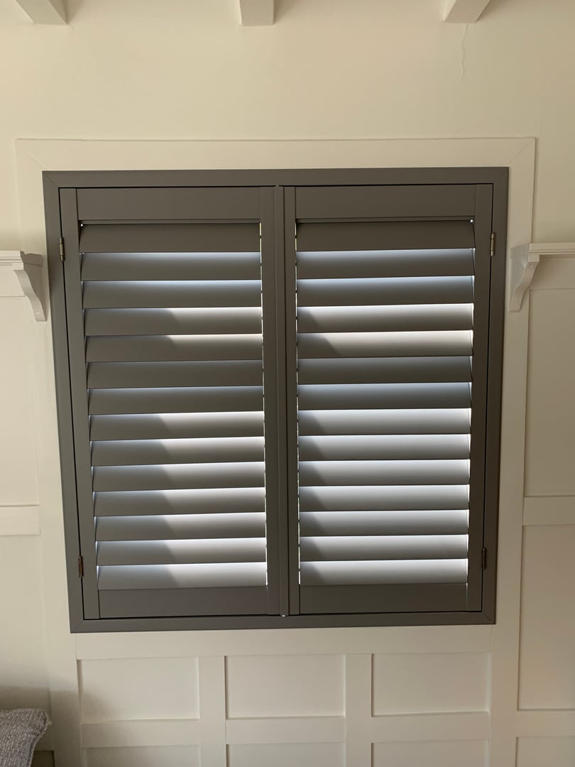 Closed gray plantation shutters on a window with white walls and decorative paneling.