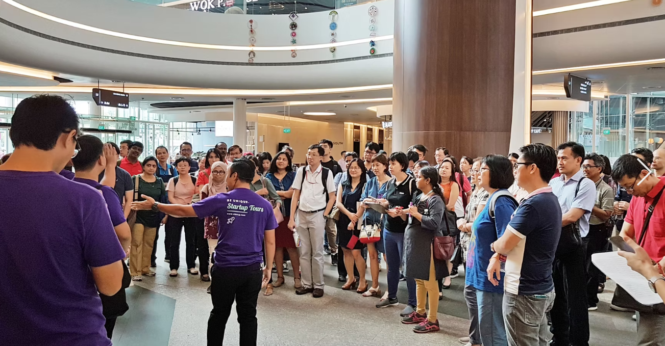 A large group of people gathered indoors listening to a speaker in a purple shirt at a public event.