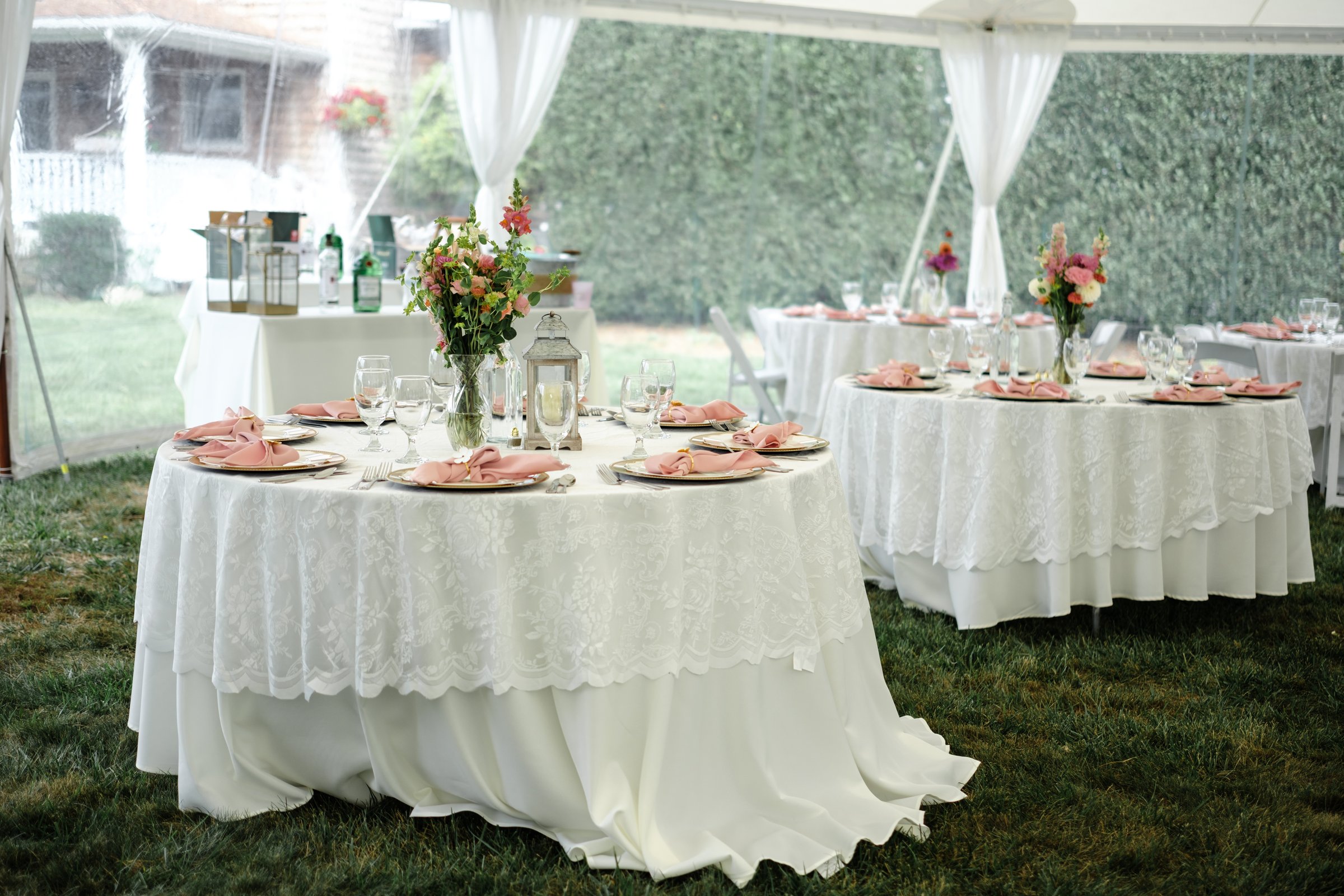 An elegant outdoor wedding reception setup featuring table  draped in crisp white tablecloths and adorned with lush floral centerpieces. The tables sit beneath a tent, creating a sophisticated and romantic atmosphere, ideal for celebrations and events.