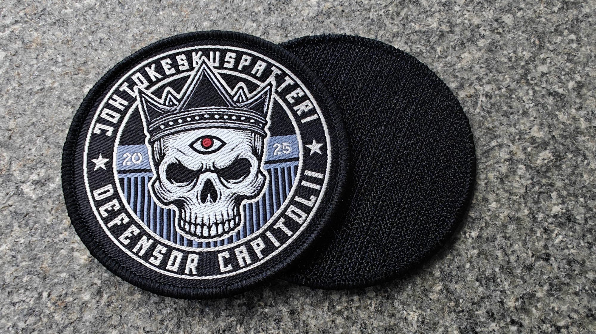 A circular patch featuring a skull with a crown and a central eye, surrounded by text and stitched in white on a dark background.