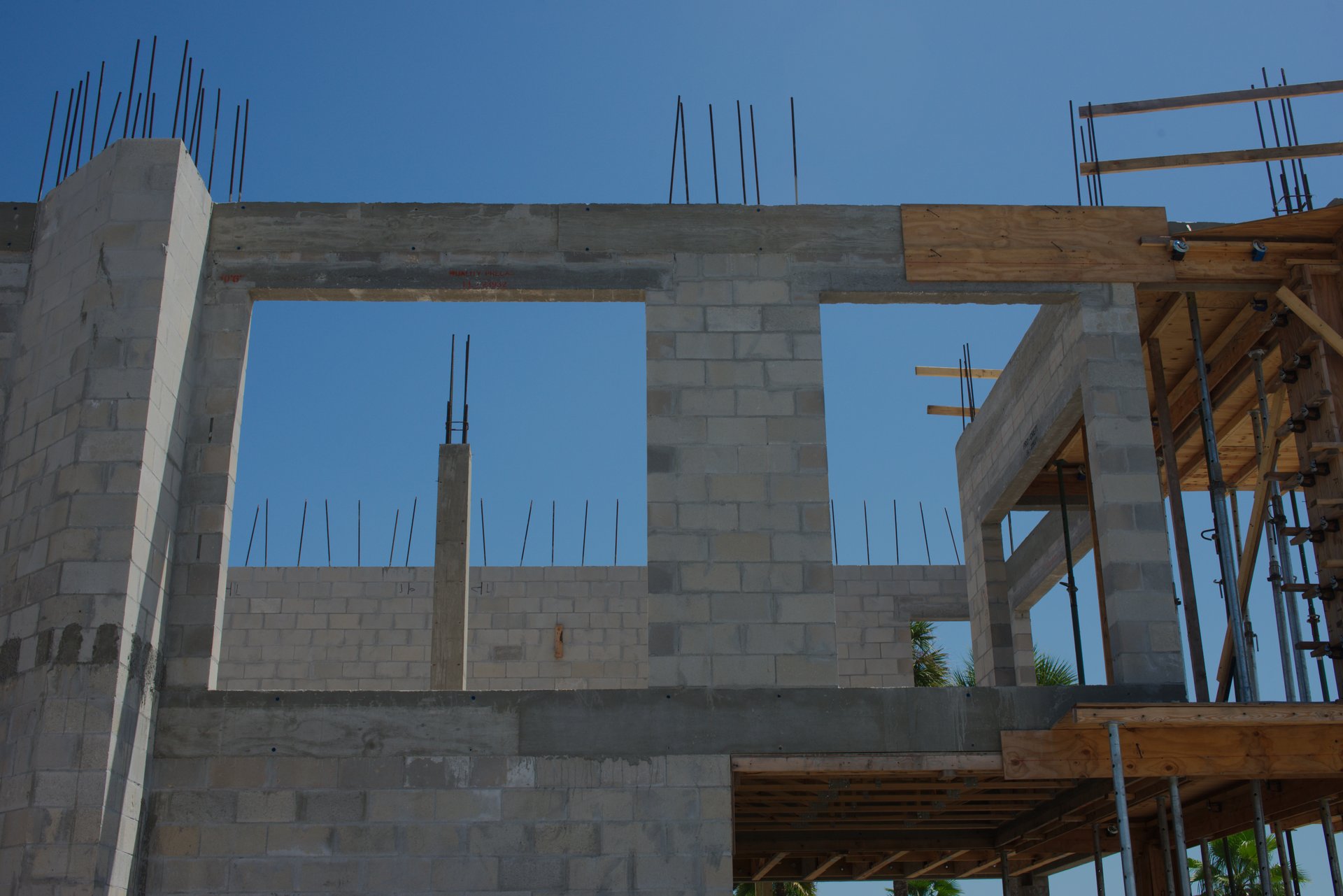 Under a clear sky, a new house structure undergoes construction, showcasing tall walls, open frames, and stacked building materials. Detailed elements highlight the construction progress and future residential development.