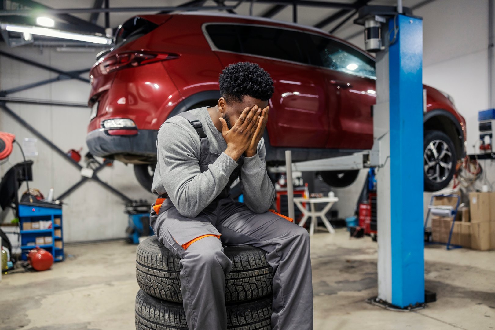 Multiracial depressed and overworked car technician sitting on pile of tires at car service and crying.