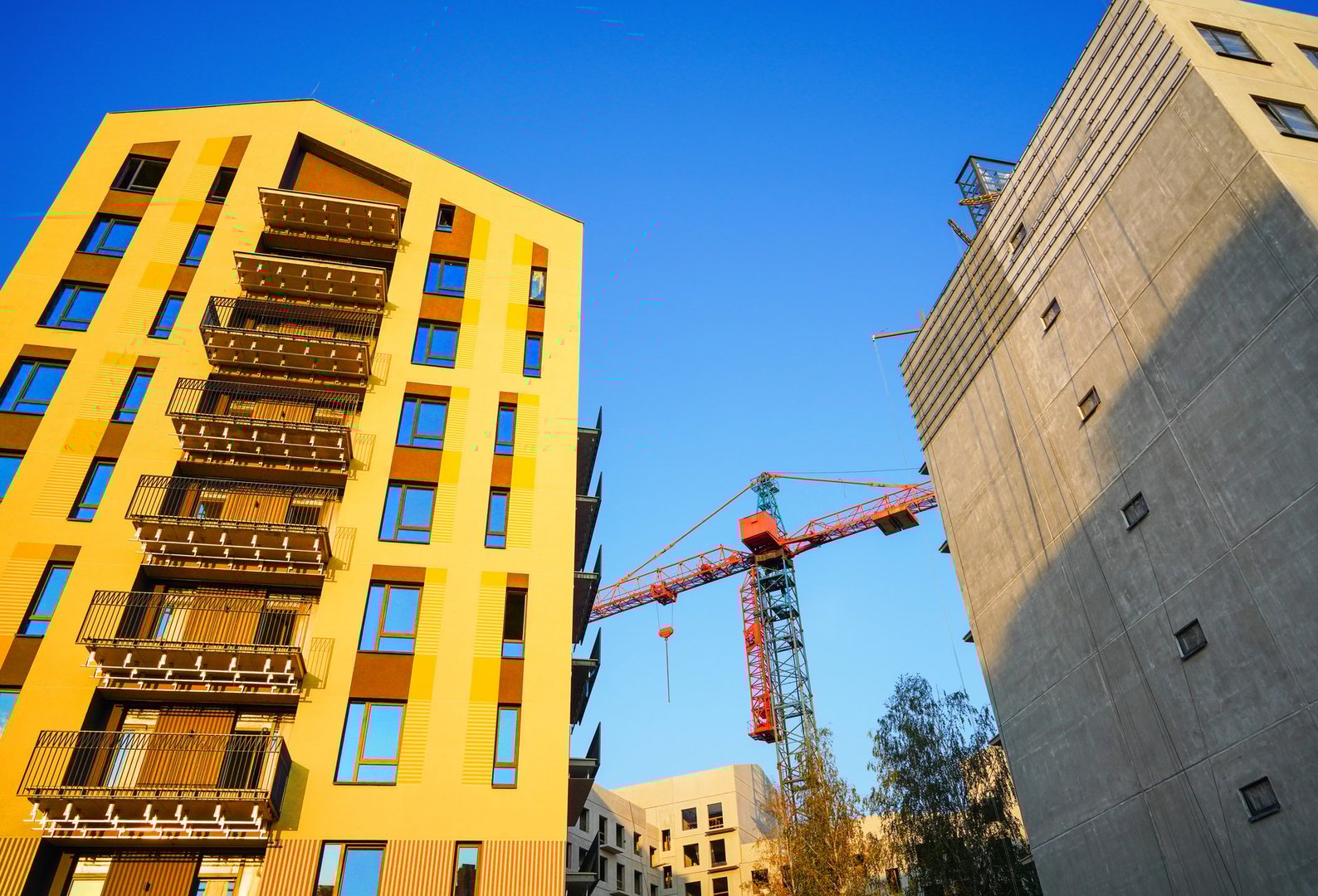 Facade of residential building in bright yellow pain. Building under construction. Modern Architecture. Vibrant yellow building onblue sky. Cranes on building construction. Newly built modern house.