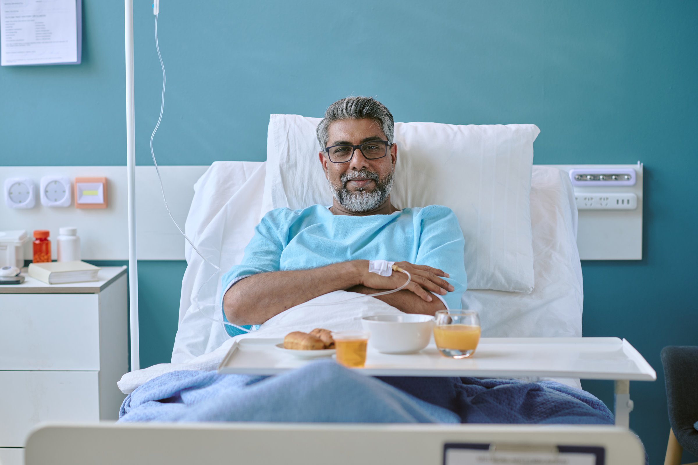 Portrait of middle aged biracial man sitting in hospital bed with IV in arm, wearing patient gown, looking at camera, having breakfast with juice and food tray on table